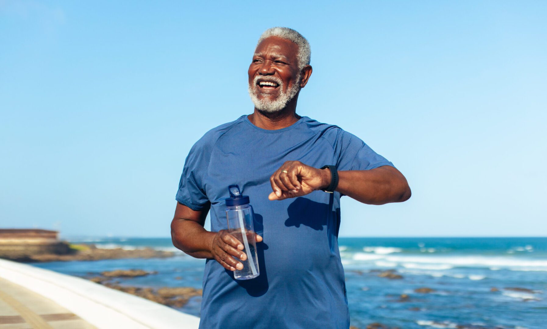 Mature man exercising at the seaside, wearing a fitness watch. He is holding a water bottle and looking enthusiastic and healthy - focusing on longevity.