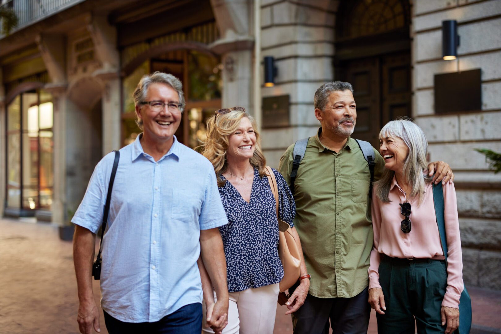 Two mature couples walking together, smiling and talking during a trip on week end. Happy elderly men and smiling beautiful women enjoy a walk in the town. Group of multiethnic seniors walking arm in arm and laughing outdoors during a trip holiday.
