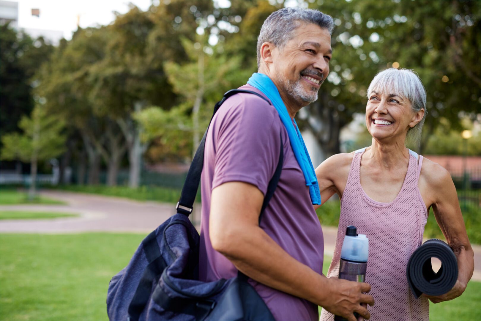 Mature multiethnic couple smiling after a fitness workout in the park. Mixed race man and elderly healthy woman with gym bags and yoga mats walking at park with copy space. Fit and cheerful olders enjoying an active lifestyle together.